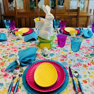 A colorful Easter table featuring vibrant plates, fuchsia and teal napkins, bright floral arrangements, and multi-colored eggs in glass jars.