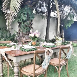 An outdoor Easter table set in a garden with floral-patterned tableware, woven baskets of fruit, potted plants, and fairy lights.