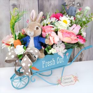 A countryside-inspired Easter centerpiece featuring a miniature wheelbarrow filled with fresh flowers, faux moss, and pastel Easter eggs.