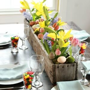 A charming farmhouse Easter centerpiece using a wooden box filled with greenery, flowers, speckled eggs, and small bunny figurines.